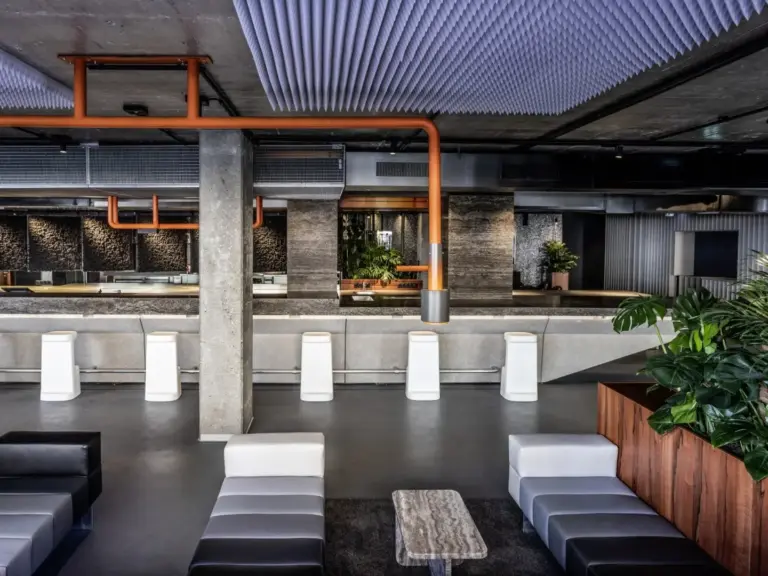 Modernes Interieur mit einer minimalistischen Bar, weißen Hockern, Betonsäulen, freiliegenden orangefarbenen Rohren, geometrischen Deckenpaneelen, schwarz-weißen Sitzgelegenheiten und Topfpflanzen, die den industriellen Raum begrünen.