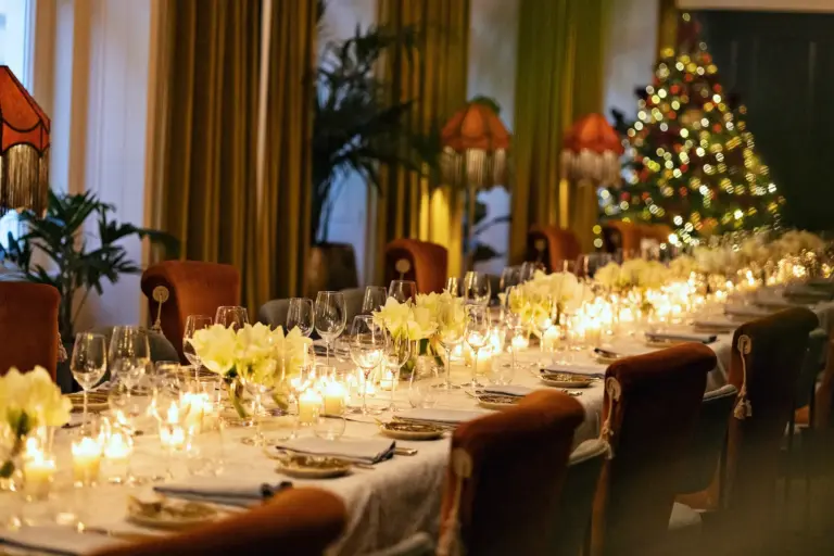 Bel Étage Hotel Motto Ein langer, eleganter Esstisch ist mit weißen Blumen, Kerzen und Gläsern gedeckt. Der Raum ist warm beleuchtet, mit Vintage-Lampen ausgestattet und im Hintergrund steht ein geschmückter Weihnachtsbaum.