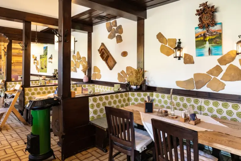 Ein gemütliches Restaurant mit Holztischen und -stühlen, grün-weiß gemusterten Fliesen an der unteren Wand, rustikalem Dekor, an der Wand befestigten Laternen und Dekorationsgegenständen an den Wänden. Natürliches Licht erhellt den Raum.