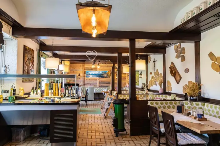 Ein gemütliches Restaurant mit Holzbalken, rustikalen Möbeln, dekorativen Lampen und ausgestellten Olivenölflaschen. Es gibt gedeckte Tische zum Essen, grün gepolsterte Bänke und Wanddekorationen mit Naturmotiven.