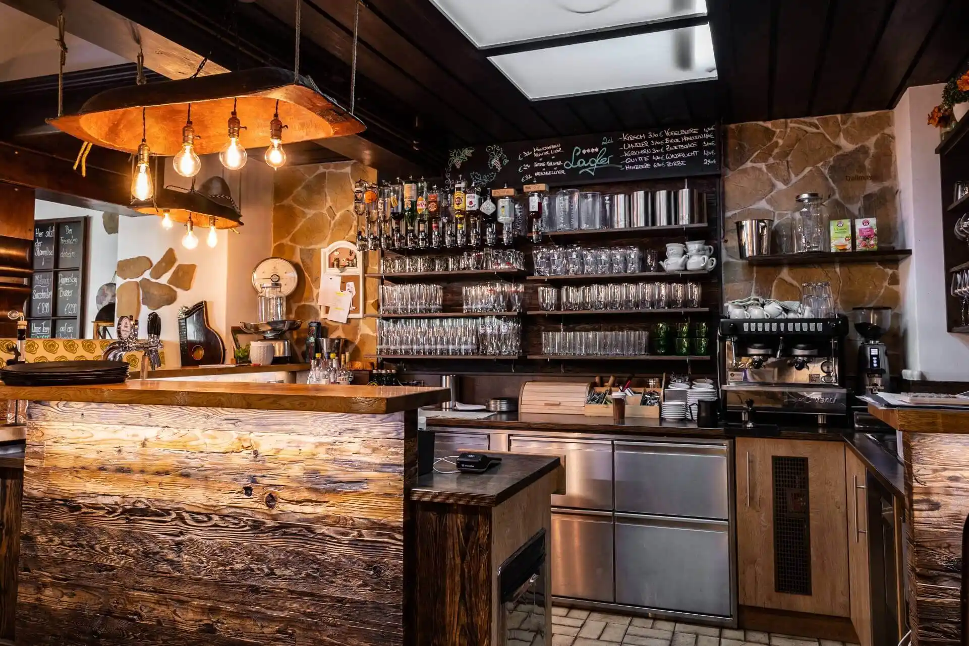 Eine gemütliche, rustikale Bar mit einem Holztresen, Regalen mit Gläsern und Flaschen, hängenden Glühbirnen, einer Kaffeemaschine und einer Speisekarte mit Kreidetafel an der Wand. Die warme Beleuchtung schafft eine einladende Atmosphäre.