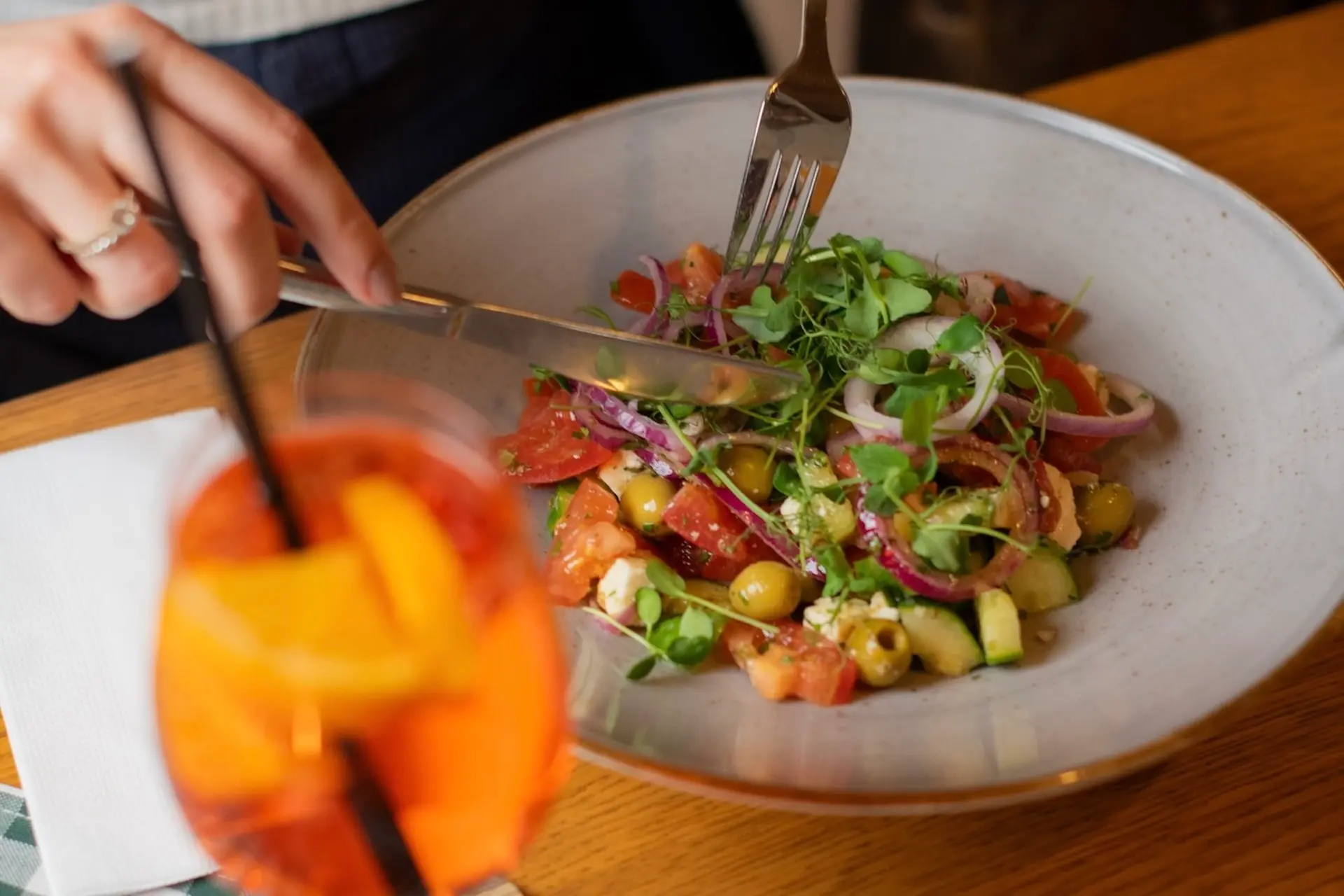 Eine Person benutzt Gabel und Messer, um einen frischen Salat mit Tomaten, Gurken, Oliven, roten Zwiebeln und Grünzeug zu essen. Im Vordergrund steht ein Glas mit einem Orangengetränk und Obstscheiben auf einem Holztisch.