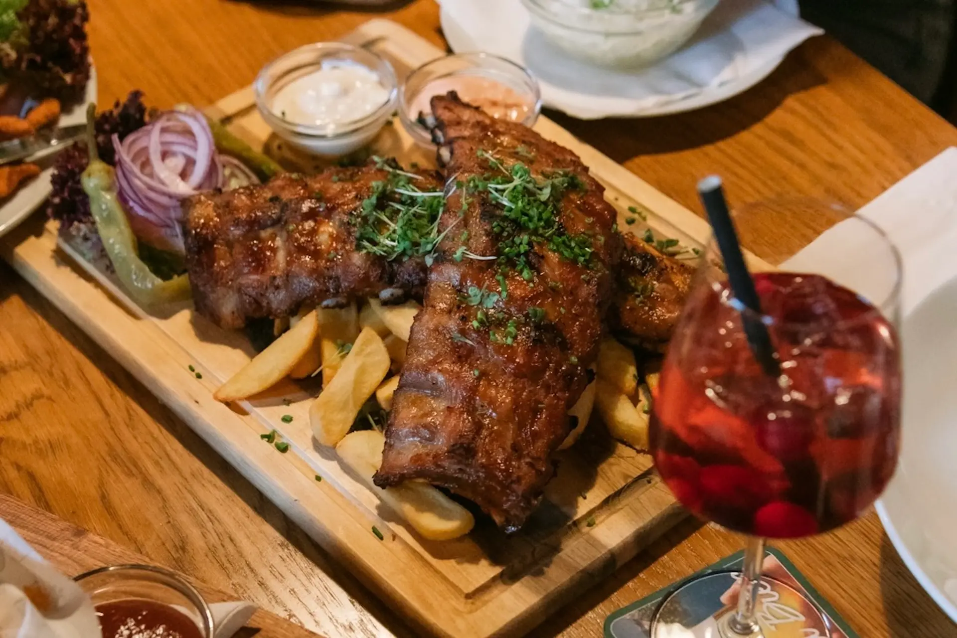 Eine Holzplatte mit gegrillten Rippchen mit frischen Kräutern, serviert mit Pommes frites, geschnittenen Zwiebeln, Zitrone, Salat und verschiedenen Dip-Saucen. Ein rotes Getränk mit Früchten und einem Strohhalm steht daneben auf dem Holztisch.