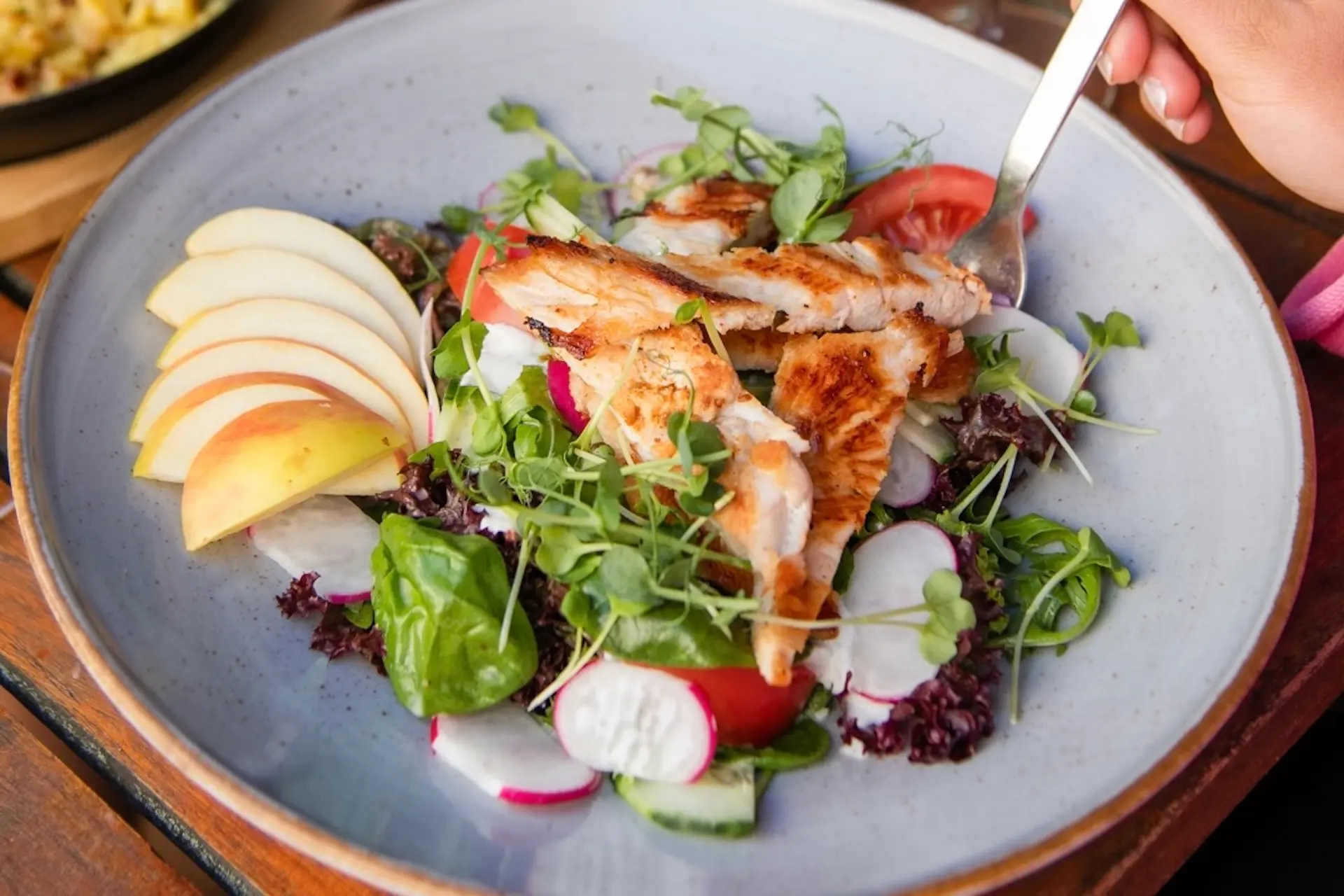 Ein Salatteller mit gegrillten Hähnchenstreifen, verschiedenen Blattsalaten, geschnittenen Radieschen, Tomatenscheiben und Apfelspalten. Eine Hand hält eine Gabel, mit der sie ein Stück Hähnchenfleisch aus dem Salat aufhebt.