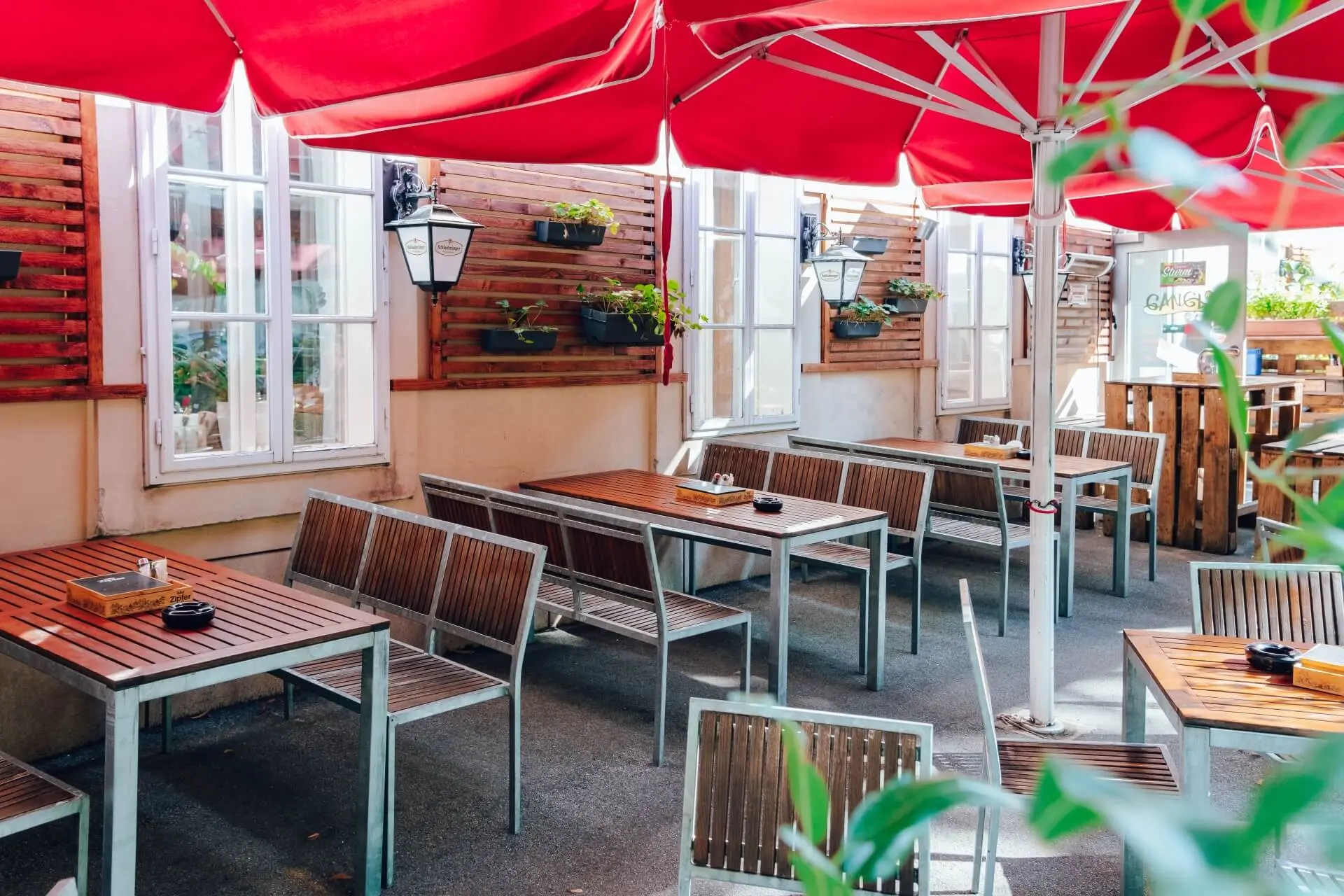 Sitzbereich im Freien mit Holztischen und -bänken unter leuchtend roten Sonnenschirmen, umgeben von weiß gerahmten Fenstern und Topfpflanzen an den Wänden. Die Szene ist leer und gut beleuchtet.