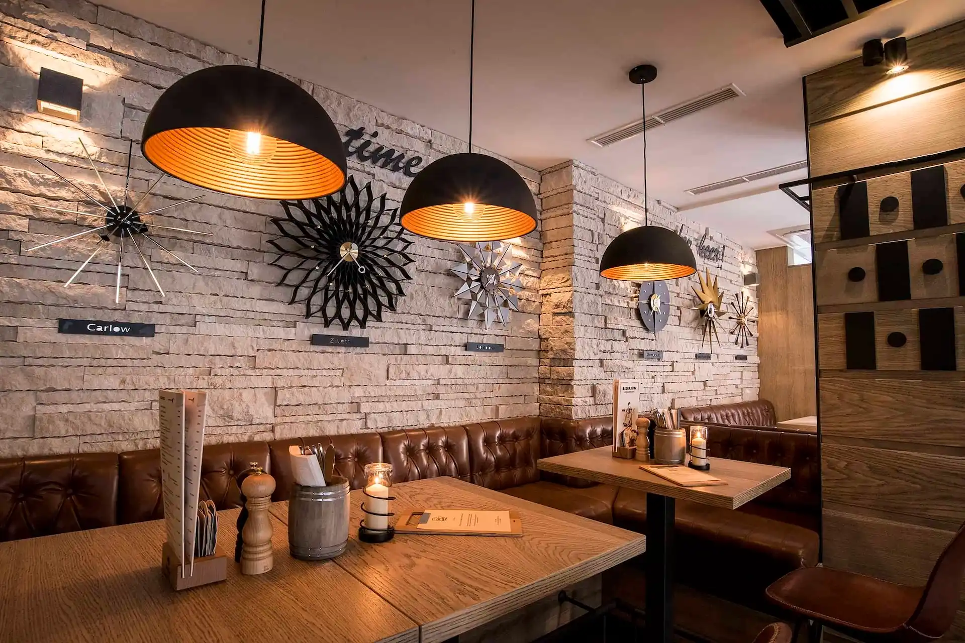 Ein gemütliches Restaurantinterieur mit braunen Leder-Sitznischen, Holztischen, hängenden Pendelleuchten und dekorativen Wanduhren an einer weißen Backsteinwand. Auf den Tischen liegen Speisekarten und Gewürze.