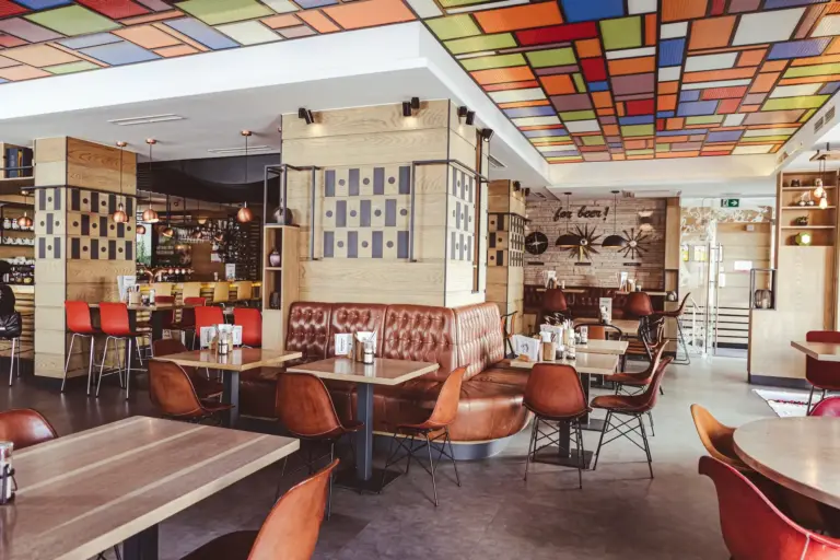 Bierraum Modernes Restaurantinterieur mit braunen Leder-Sitznischen, Holztischen, orangefarbenen Stühlen und einem farbenfrohen, geometrischen Deckenmuster. Der Raum ist hell beleuchtet und strahlt eine gemütliche, moderne Atmosphäre aus.
