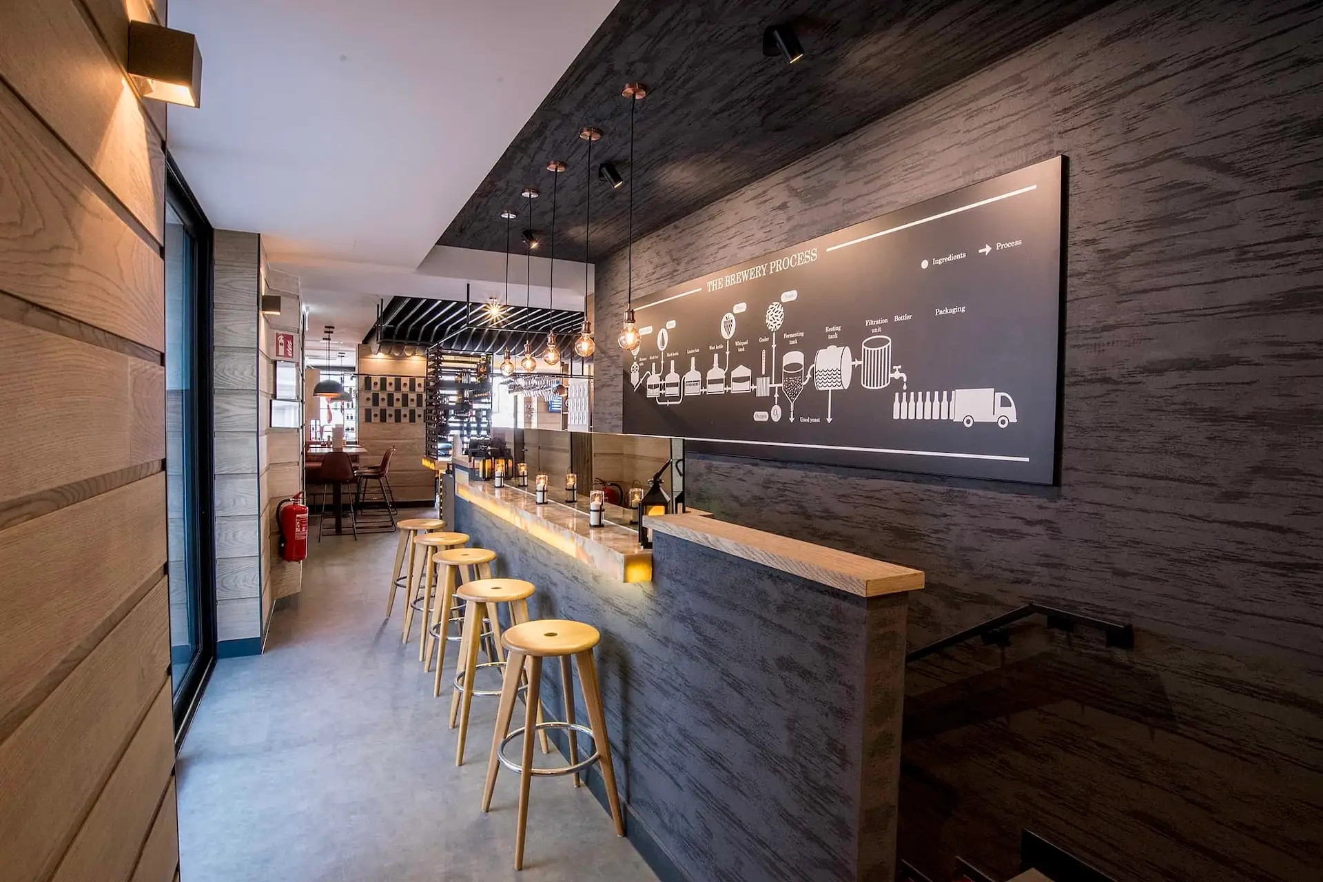 Eine moderne Café-Einrichtung mit Holzhockern an der Theke, warmer Beleuchtung und einer großen Darstellung des Brühprozesses an einer dunklen, strukturierten Wand. Im Hintergrund sind Tische und Stühle in der Nähe großer Fenster zu sehen.