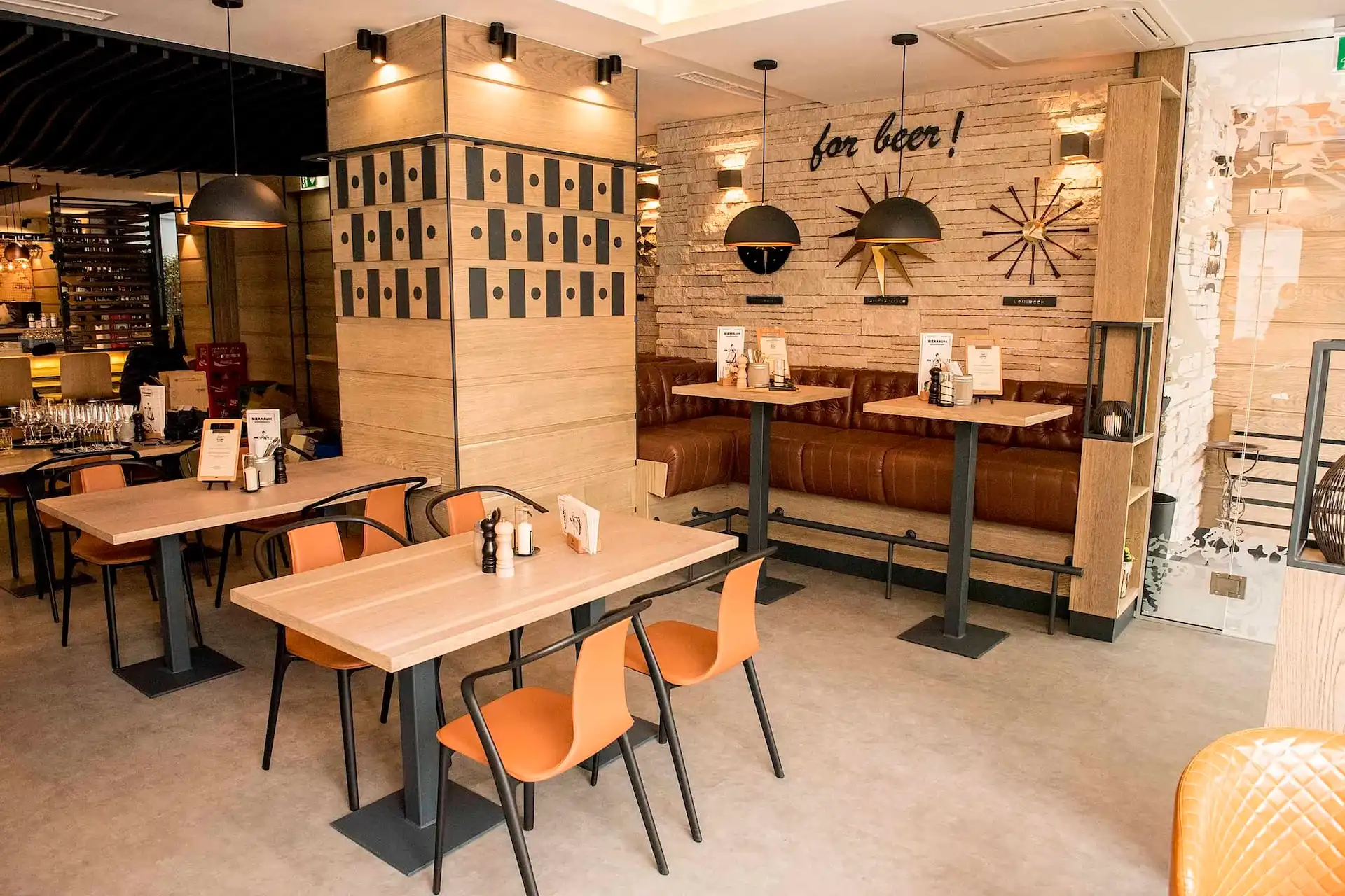 Moderne Restaurantausstattung mit hellen Holztischen, orangefarbenen Stühlen, braunen Leder-Sitznischen und dekorativen Wandbildern; Speisekarten und Gewürze liegen auf den Tischen, und der Raum ist hell und aufgeräumt.