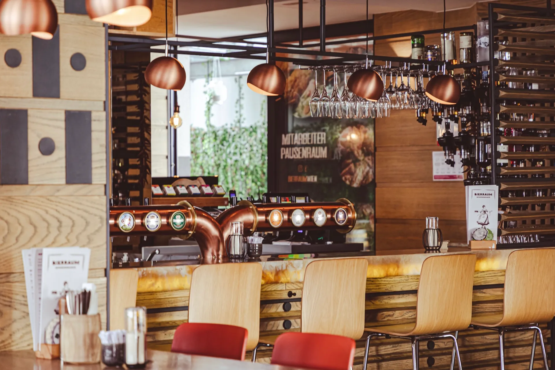 Eine moderne Bar mit Holzdekor, Bierhähnen, hängenden Gläsern und hohen Stühlen. Warme Beleuchtung und Grünpflanzen sorgen für eine gemütliche Atmosphäre.