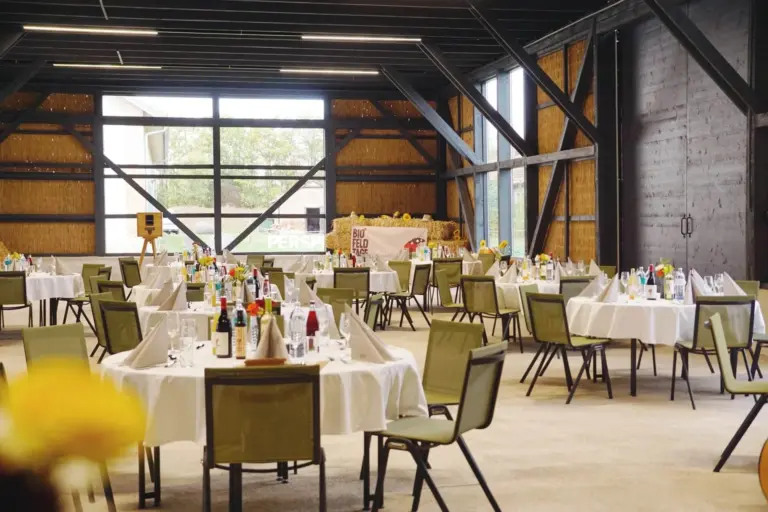 Ein geräumiger Veranstaltungssaal mit runden Tischen, die mit weißen Tischdecken bedeckt und mit Weinflaschen, Gläsern und Servietten gedeckt sind. Grüne Stühle umgeben die Tische, und große Fenster lassen natürliches Licht herein.