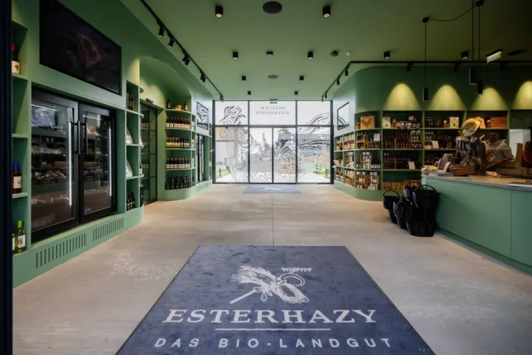 Innenraum eines modernen Bioladens mit grünen Wänden, Regalen mit verschiedenen Produkten, einer großen Bodenmatte mit der Aufschrift Esterhazy Das Bio-Landgut und großen Glastüren am Eingang.