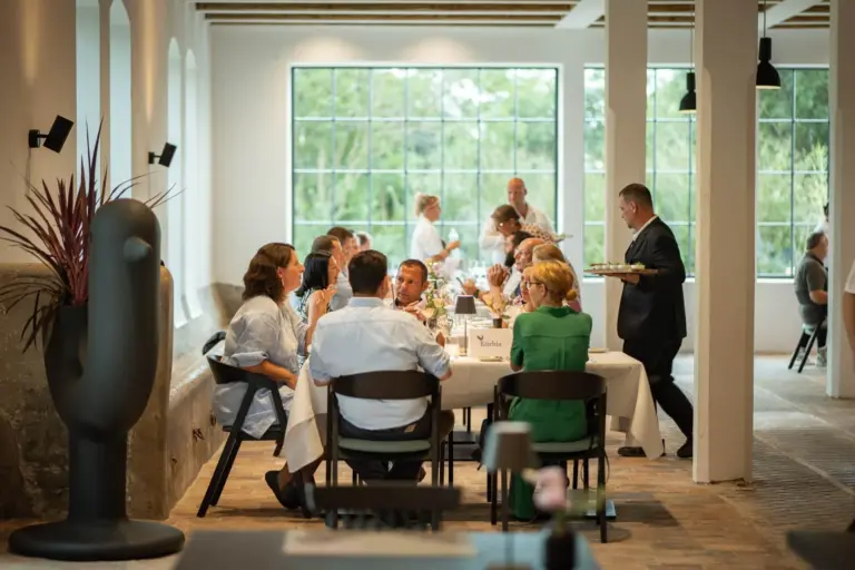 Eine Gruppe von Menschen sitzt um einen Tisch herum und genießt eine Mahlzeit in einem hellen, modernen Restaurant mit großen Fenstern. Ein Kellner steht in der Nähe und serviert Getränke, während sich andere an Tischen im Hintergrund unterhalten.