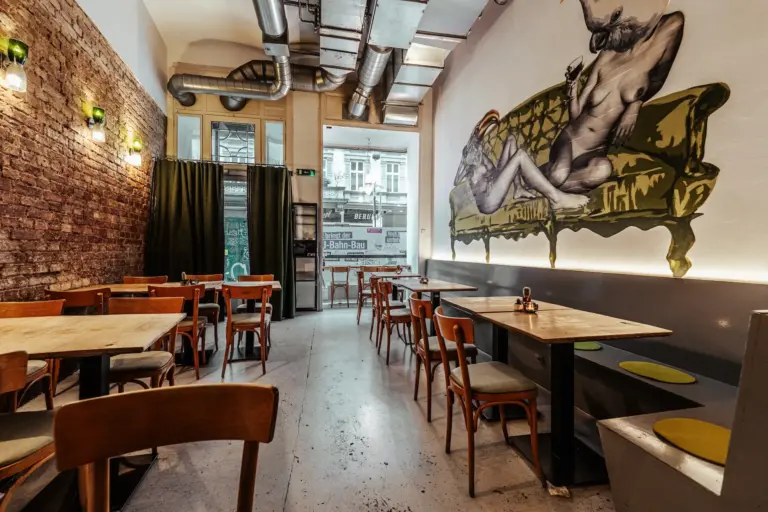 Ein gemütliches Café mit Holztischen und -stühlen, sichtbaren Ziegelwänden, einer industriellen Decke und einem großen Wandgemälde mit einer liegenden Figur auf einer grünen Couch. Durch die vordere Glastür und die Fenster fällt natürliches Licht ein.