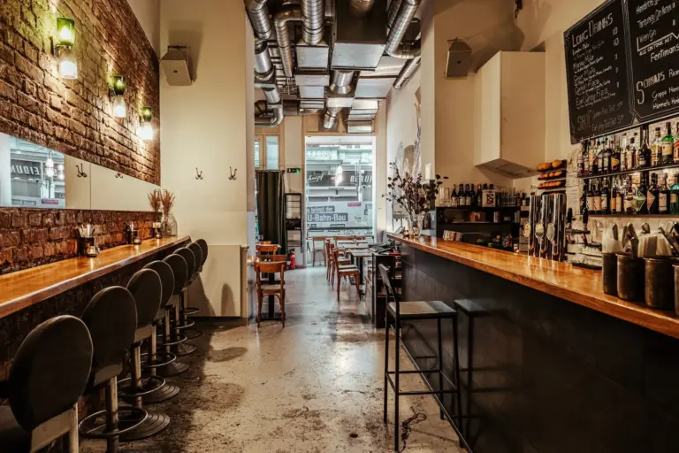Eine gemütliche, moderne Bar mit langem Tresen, hohen Hockern, sichtbaren Ziegelwänden, industrieller Decke und kleinen Tischen, die zu einem Fenster im hinteren Bereich führen. Regale mit Flaschen und Speisekarten auf Kreidetafeln schmücken den Raum.