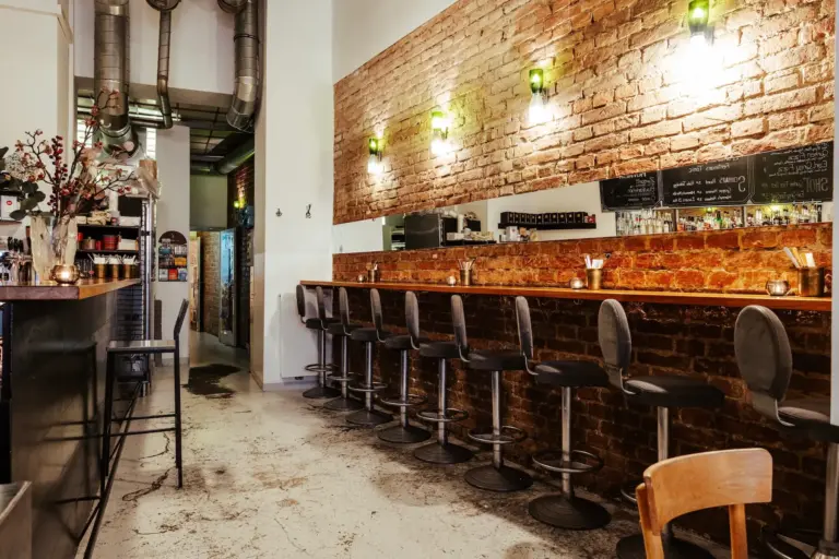 Bizú Eine gemütliche Bar mit freiliegenden Backsteinwänden, einem langen Holztresen mit Barhockern, warmer Beleuchtung und industriellen Deckenrohren. Hinter der Bar befinden sich Regale mit Flaschen und Gläsern, die eine einladende Atmosphäre schaffen.