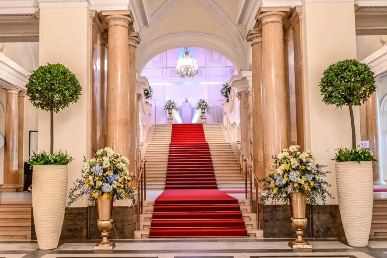 Eine große Treppe mit rotem Teppich, flankiert von hohen Säulen und großen Blumenarrangements in eleganten Vasen, führt hinauf zu einem von Kronleuchtern beleuchteten Treppenabsatz in einem luxuriösen Interieur.