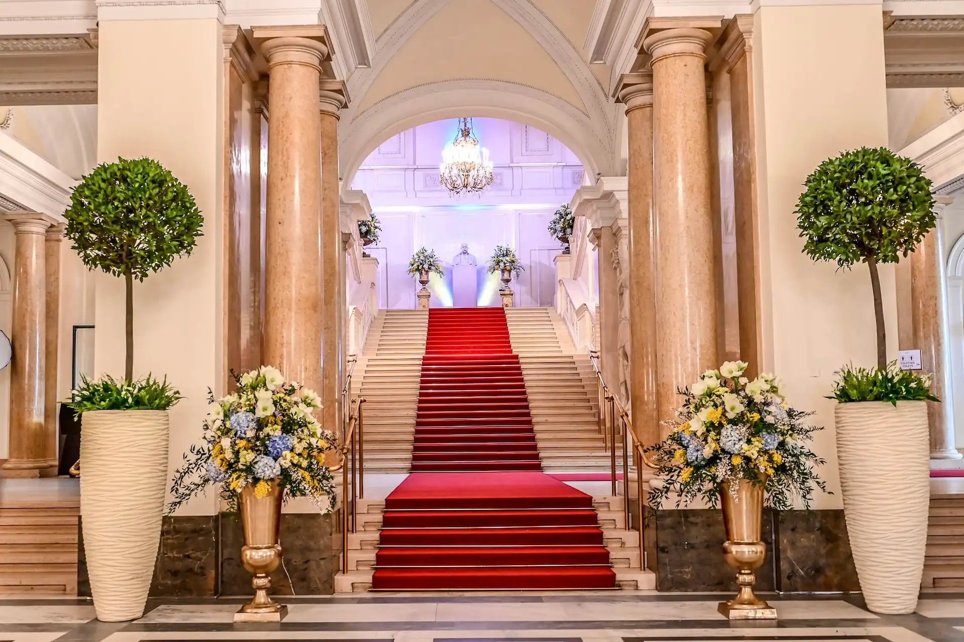 Eine große Treppe mit rotem Teppich, flankiert von hohen Säulen und großen Blumenarrangements in eleganten Vasen, führt hinauf zu einem von Kronleuchtern beleuchteten Treppenabsatz in einem luxuriösen Interieur.