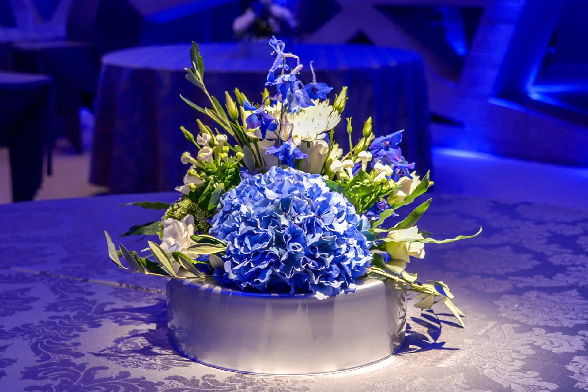 Ein Tischschmuck aus blauen und weißen Blumen, darunter Hortensien, arrangiert in einem runden Gefäß auf einem Tisch mit einer blau gemusterten Tischdecke unter blauer Stimmungsbeleuchtung.