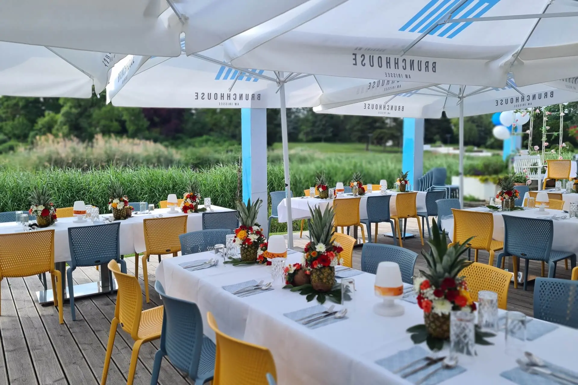 Essbereich im Freien mit gedeckten Tischen, dekoriert mit weißen Tischtüchern, bunten Blumen und Ananas, gelben und blauen Stühlen und großen weißen Sonnenschirmen, mit Blick auf eine grüne, natürliche Landschaft.