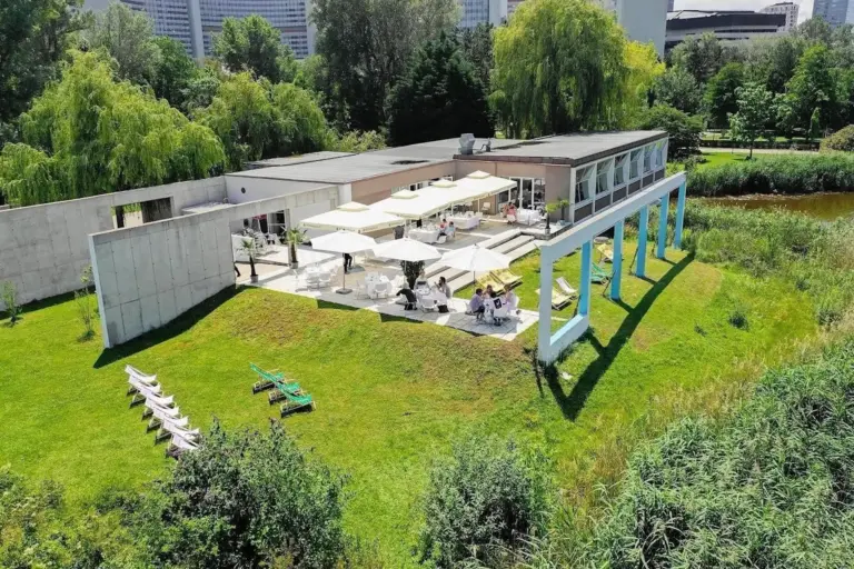 Auf einem von Bäumen umgebenen Grashügel steht ein modernes Gebäude mit großen Fenstern und einem Innenhof mit Sonnenschirmen. Die Menschen sitzen im Freien, und auf dem Rasen sind Liegestühle aufgestellt. Im Hintergrund sind hohe Gebäude zu sehen.
