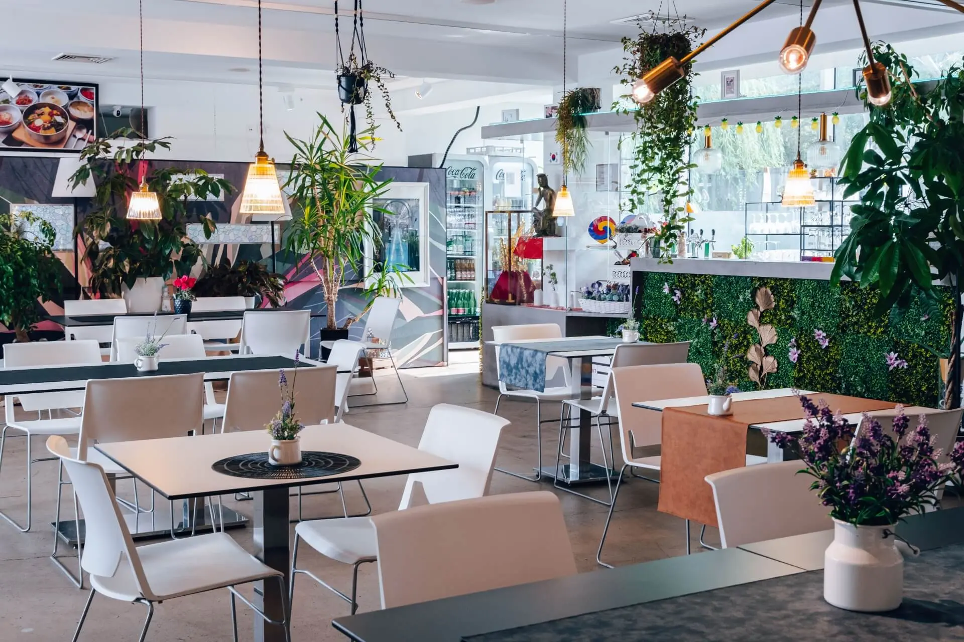 Ein modernes Café mit weißen Stühlen und Tischen, Grünpflanzen, Hängelampen und großen Fenstern, die natürliches Licht hereinlassen. Der Raum wirkt hell, sauber und einladend, mit einem Thekenbereich im Hintergrund.