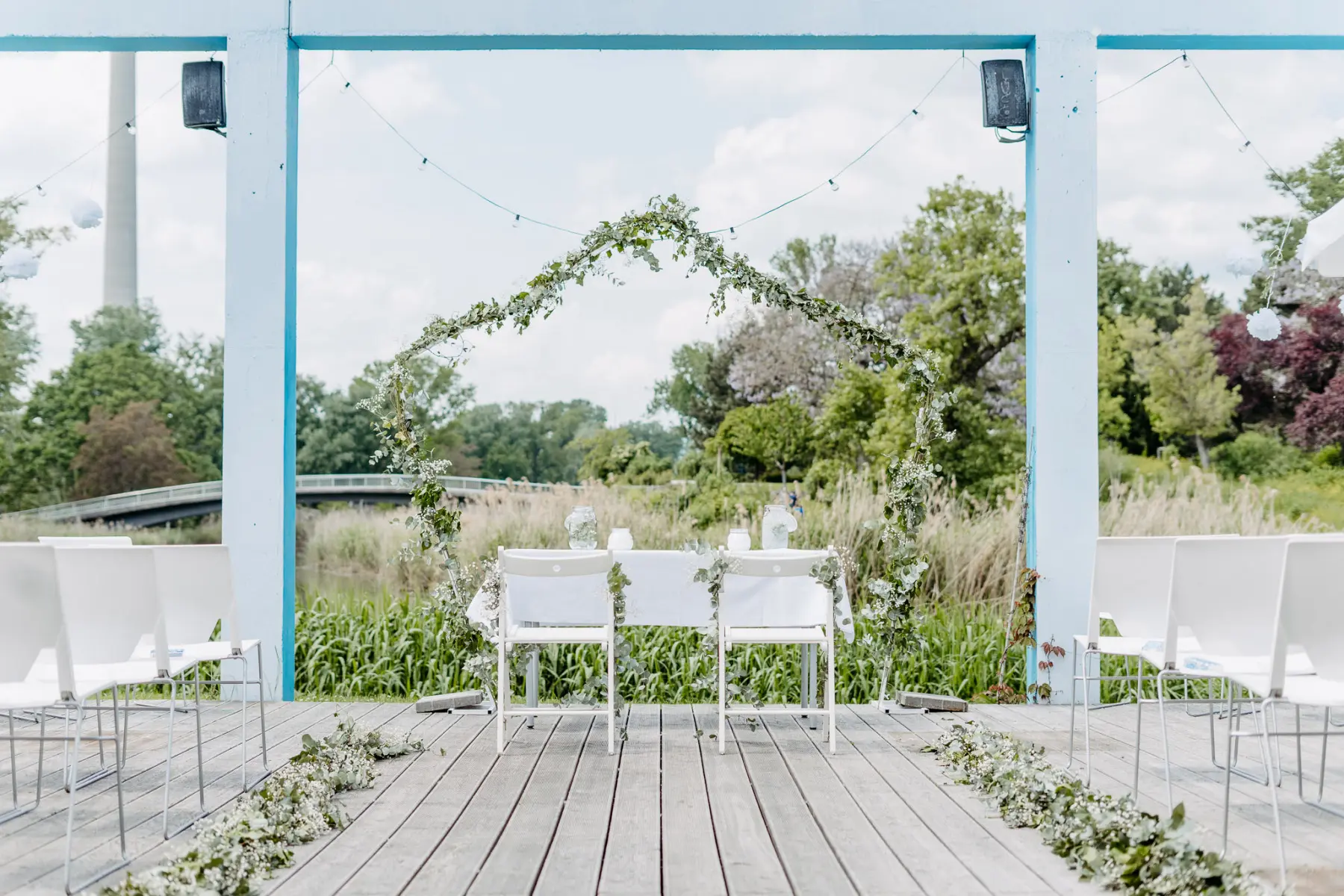 Eine Hochzeit im Freien mit zwei weißen Stühlen und einem Tisch unter einem Blumenbogen auf einer Holzterrasse, umgeben von Grün und Reihen von weißen Stühlen für die Gäste.