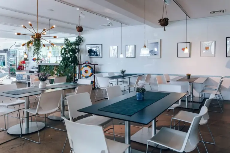 Modernes Café-Interieur mit weißen Stühlen und dunklen Tischen, dekoriert mit Hängepflanzen, gerahmten Bildern an den Wänden und Pendelleuchten, die eine helle und einladende Atmosphäre schaffen.