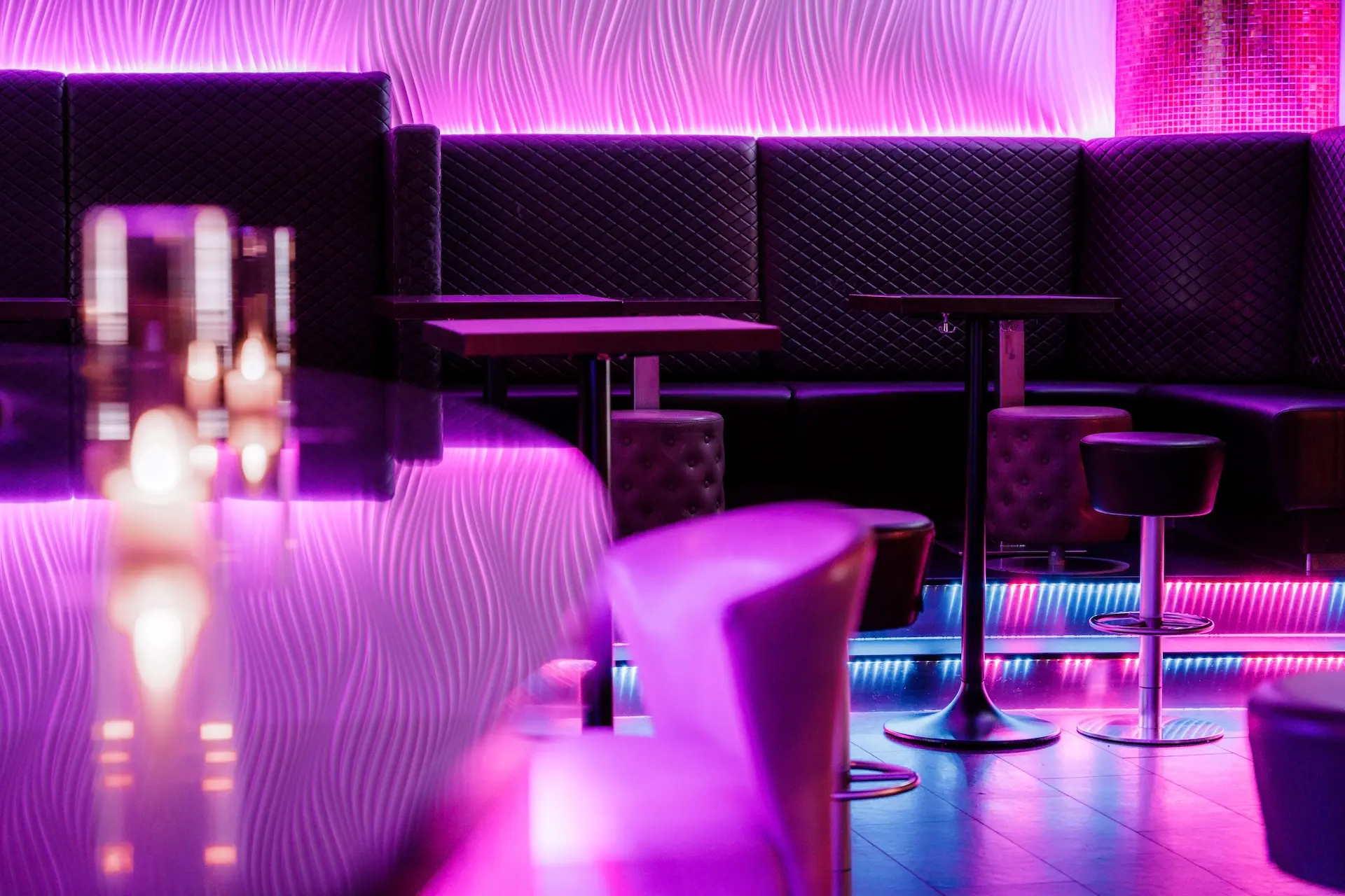 Eine moderne Lounge mit schwarz gesteppten Sitzmöbeln, kleinen Tischen und Hockern, die von lila und rosa Neonröhren beleuchtet werden. Der Bartresen spiegelt die lebendige Beleuchtung wider und schafft eine stilvolle und bunte Atmosphäre.