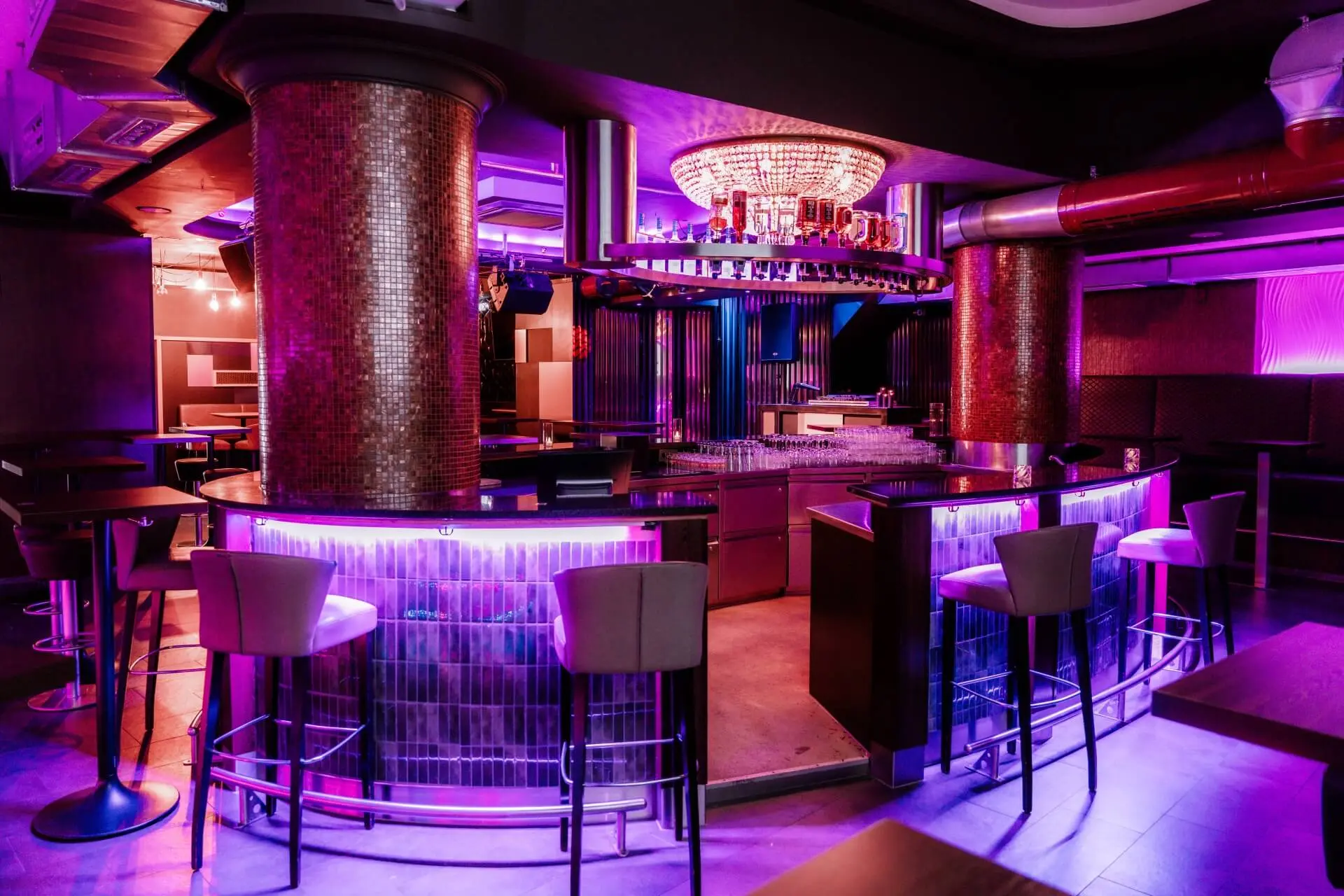 Eine moderne Bar mit lilafarbener und rosafarbener Beleuchtung, hohen Hockern rund um den Tresen, zwei mosaikgefliesten Säulen und einem Kronleuchter über dem runden Barbereich. Die Atmosphäre ist stilvoll und einladend.