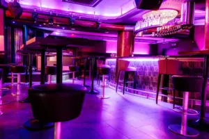 Ein moderner Nachtclub mit violetter Stimmungsbeleuchtung, hohen Tischen und Barhockern, einem glänzenden Bartresen und einem von der Decke hängenden Kronleuchter. Der Raum ist leer und lebendig, mit einem stilvollen, zeitgenössischen Design.