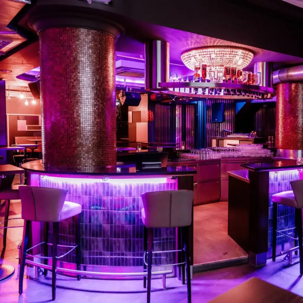 Ein stilvolles Nachtclub-Interieur mit leuchtenden violetten und rosafarbenen Lichtern, Mosaiksäulen und einer runden Bar mit hohen Stühlen - die Café Bar Laby verströmt eine moderne, lebendige Atmosphäre unter einem funkelnden Kronleuchter über dem Kopf.