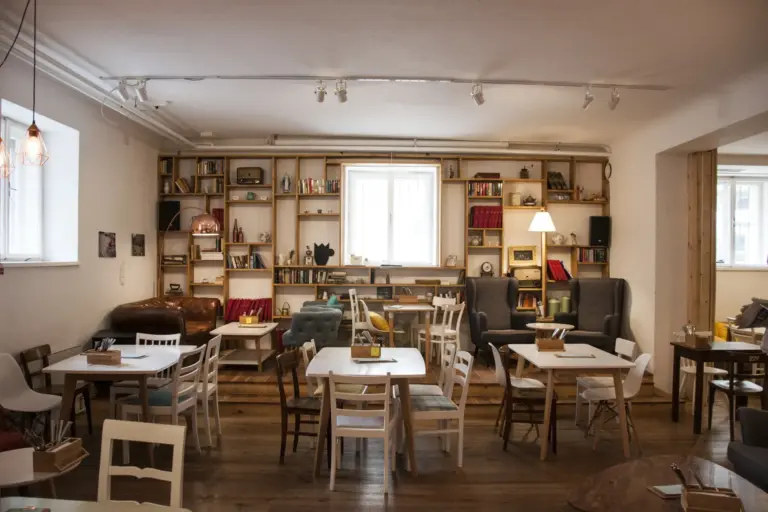 Das Café Caspar ist ein gemütliches Café mit Holzböden, uneinheitlichen Stühlen und Tischen, mit Büchern und Dekoration gefüllten Regalen und großen Fenstern, die Tageslicht hereinlassen. Der Raum hat eine warme, einladende Atmosphäre.