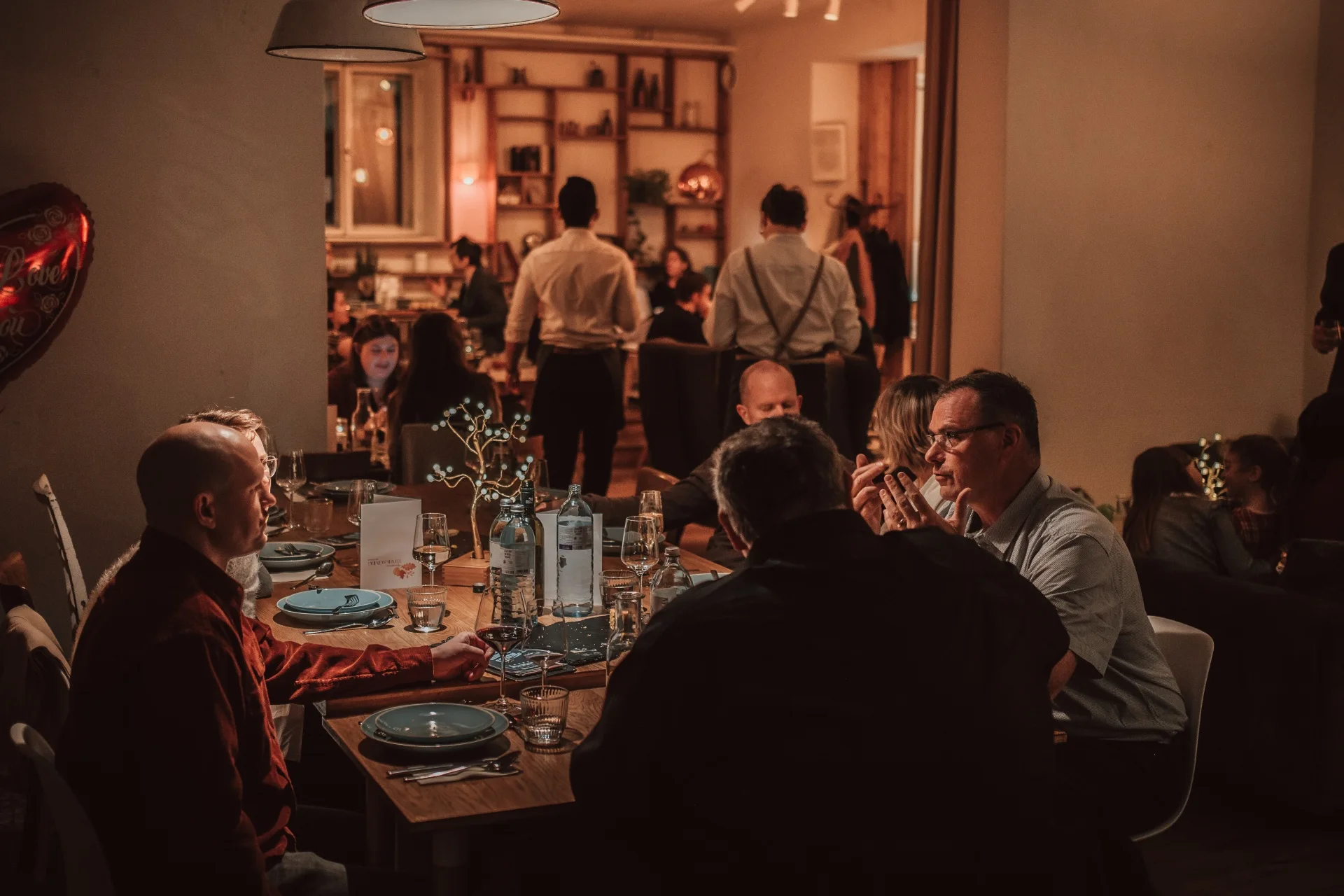 Eine Gruppe von Menschen sitzt um einen Tisch im warm beleuchteten Café Caspar und unterhält sich. Im Hintergrund sind weitere Gäste und das Restaurantpersonal zu sehen, die eine gemütliche, lebhafte Atmosphäre schaffen.