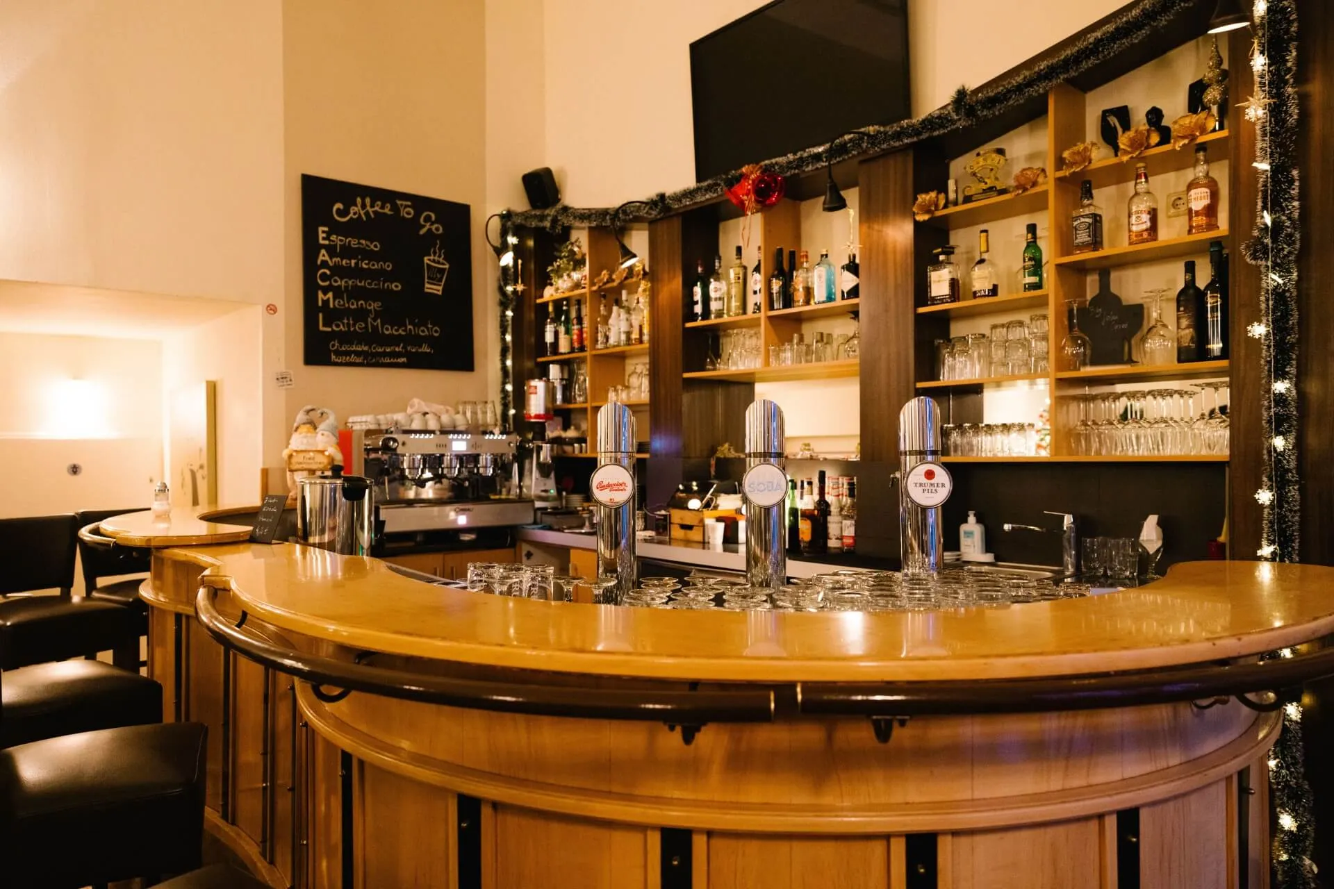 Eine gemütliche Bar mit einem hölzernen Tresen, drei Bierzapfhähnen, Regalen mit Schnapsflaschen und Gläsern, einer Speisekarte auf Kreidetafeln, Kaffeemaschinen und warmer Beleuchtung, die eine einladende Atmosphäre schafft.