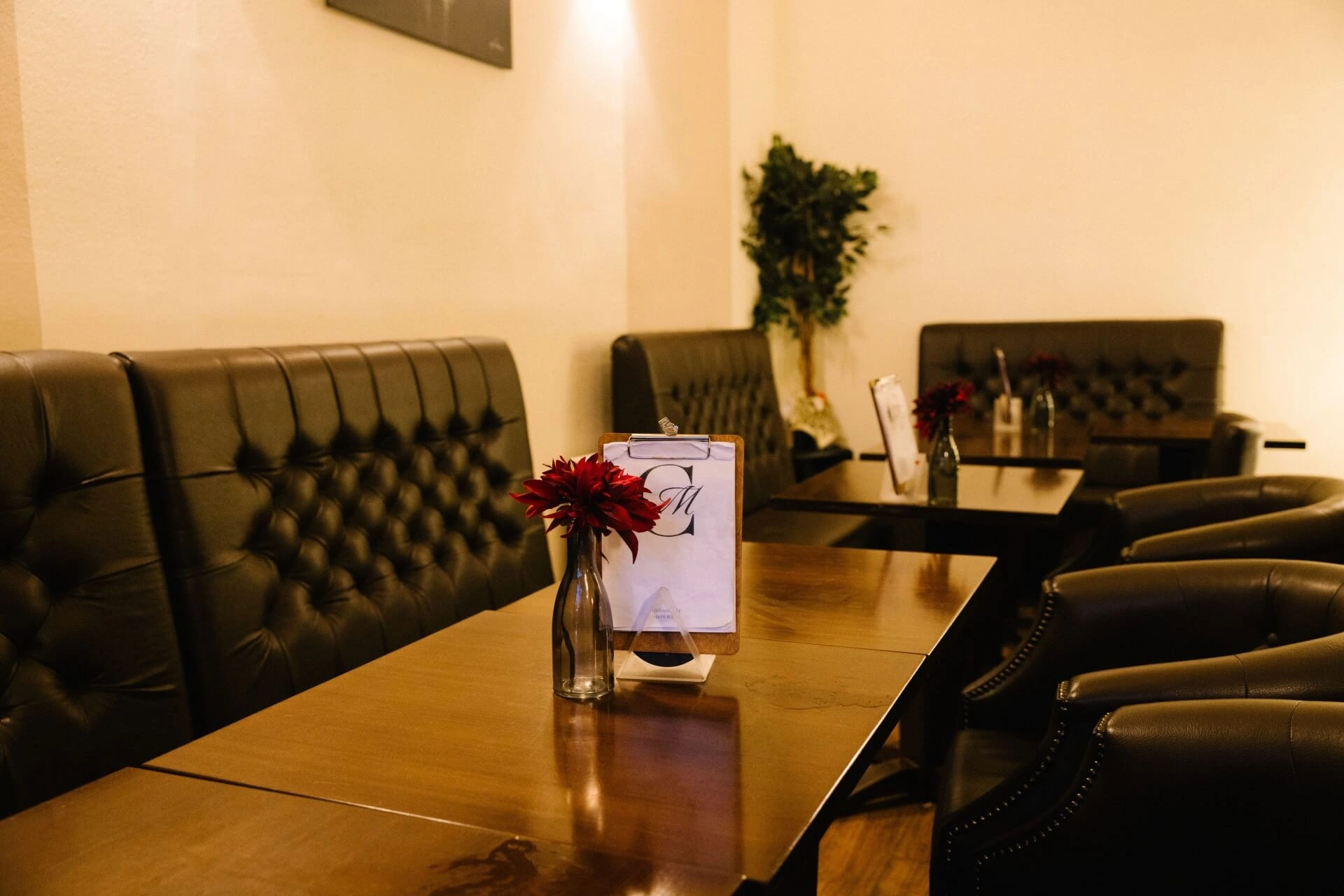 Eine gemütliche Café-Einrichtung mit dunklen Ledersesseln, Holztischen, kleinen Vasen mit roten Blumen und einer Speisekarte auf einem Ständer. Warme Beleuchtung und eine Topfpflanze in der Ecke schaffen eine einladende Atmosphäre.