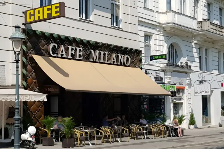 Straßenansicht des Cafe Milano mit Sitzplätzen im Freien und Menschen, die an Tischen unter einer großen braunen Markise sitzen. Das Café befindet sich im Erdgeschoss eines weißen Gebäudes, und in der Nähe sind Geschäfte und Schilder zu sehen.