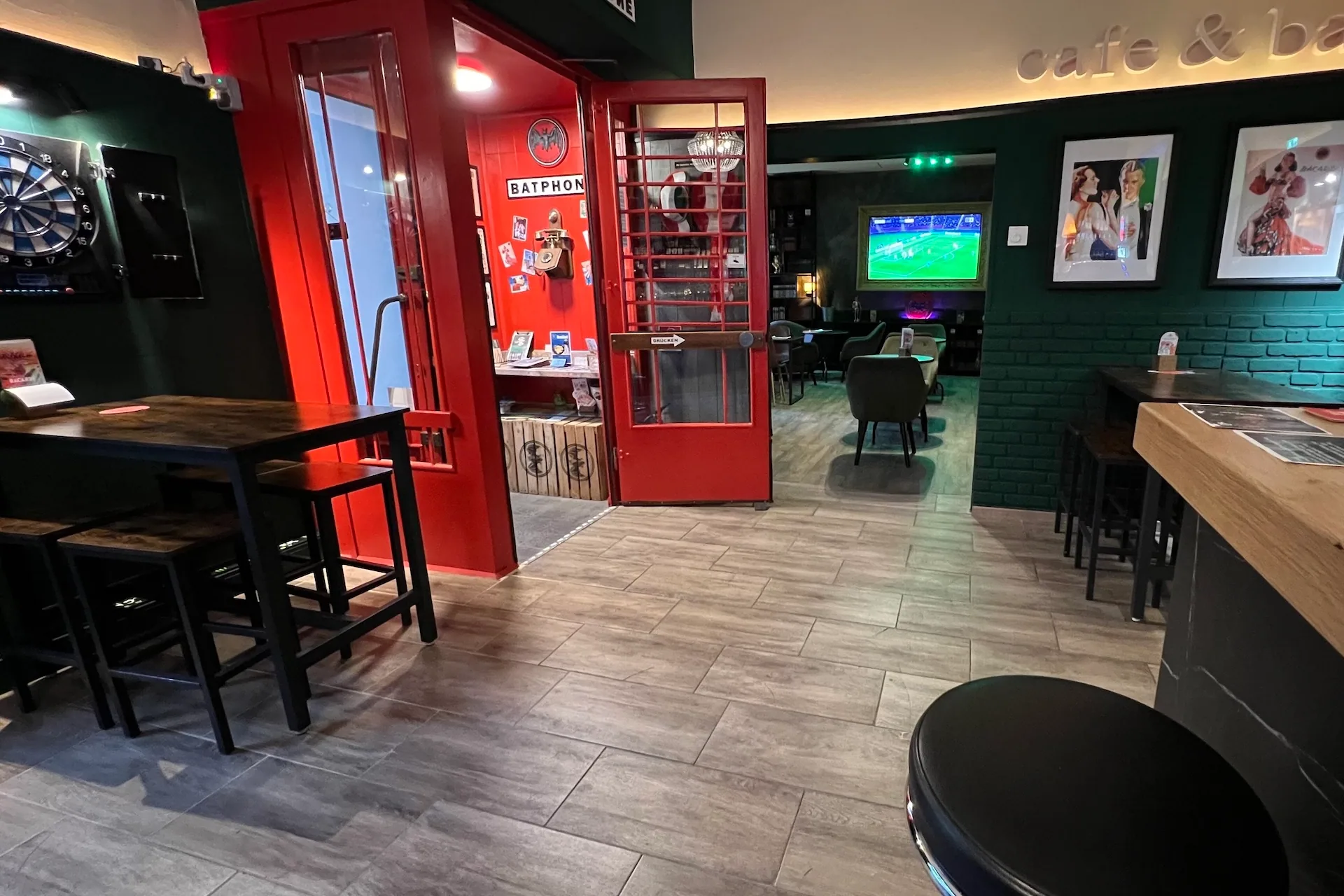 Eine gemütliche Bar mit Holztischen und Hockern, einer Dartscheibe an der Wand und einer roten Telefonzelle. Im Hintergrund sehen Leute ein Fußballspiel auf einem großen Fernseher. Gerahmte Fotos hängen an einer dunkelgrünen Wand.