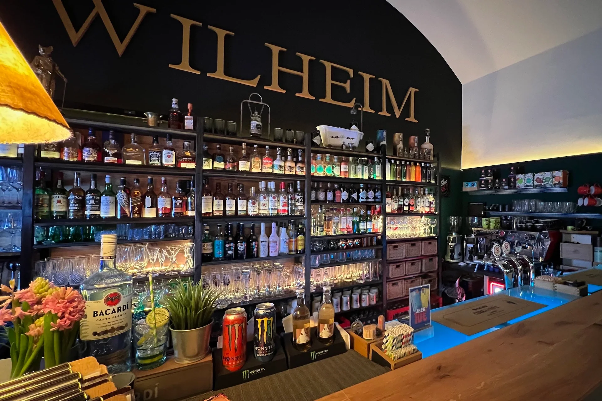 Eine gut bestückte Bar mit Regalen mit Schnapsflaschen, Gläsern und Pflanzen hinter einem Holztresen. Die großen goldenen Buchstaben "WILHEIM" stehen über den Regalen an einer schwarzen Wand. Die Bar ist warm beleuchtet.
