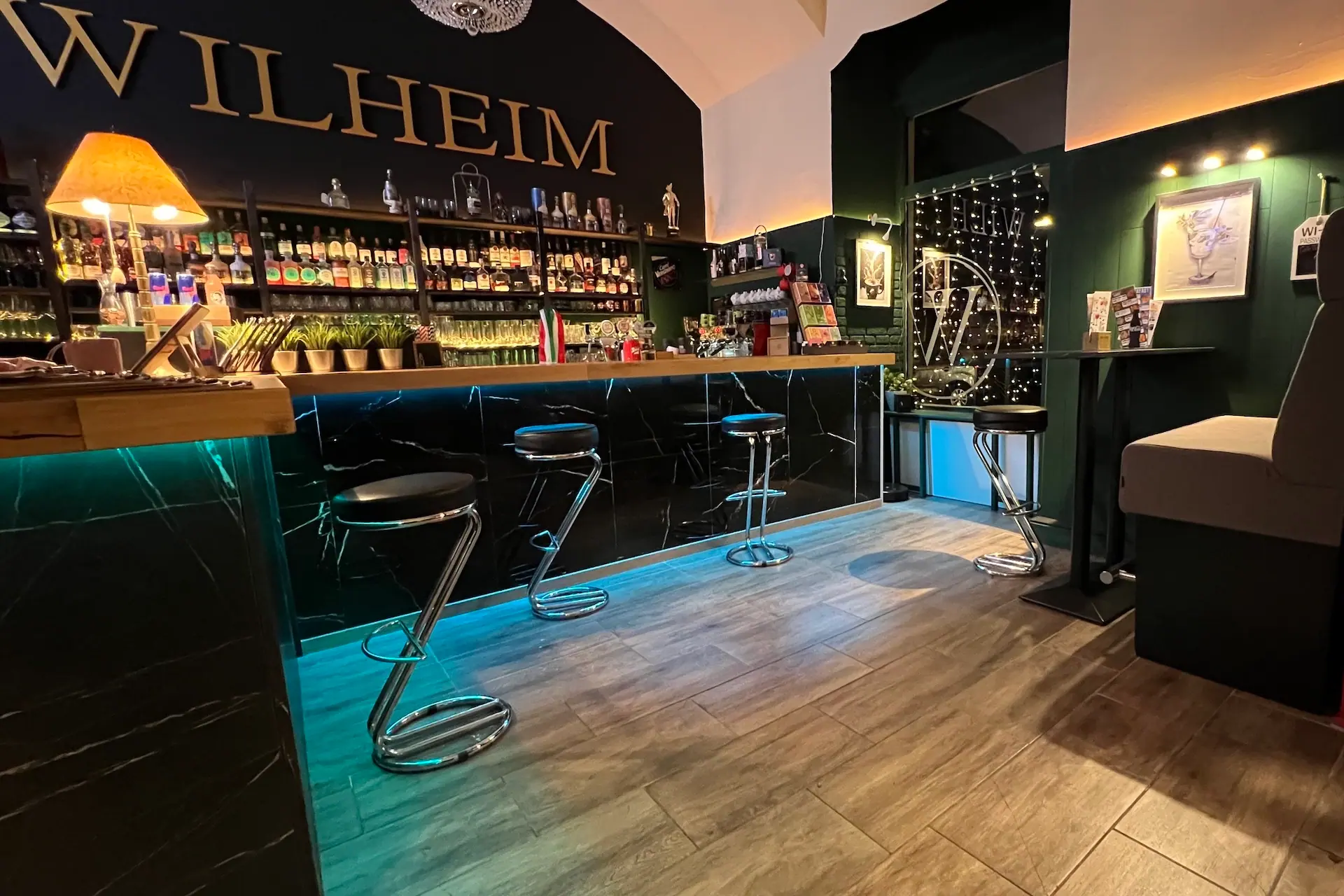Eine moderne Bar mit einem schwarzen Tresen, vier verchromten und schwarzen Hockern, die mit blauer Beleuchtung beleuchtet werden, und Regalen mit verschiedenen Flaschen. Das Wort WILHEIM prangt in großen goldenen Buchstaben an der Rückwand.