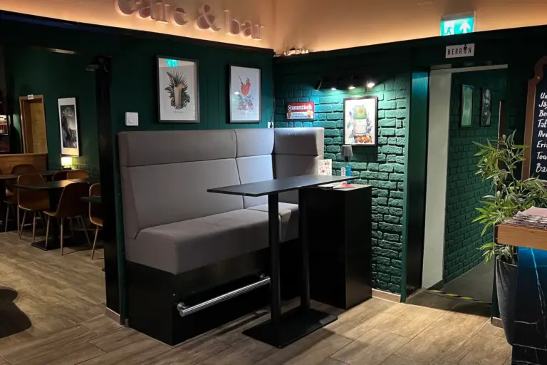 Ein gemütliches Café-Interieur mit einer grauen gepolsterten Bank, einem schwarzen Tisch, grünen Backsteinwänden, gerahmten Kunstwerken und warmer Beleuchtung; im Hintergrund sind eine Tür und ein Schild mit einer Herrentoilette zu sehen.