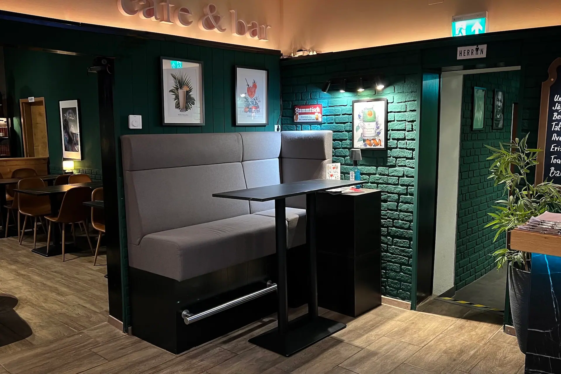 Ein gemütliches Café-Interieur mit einer grauen gepolsterten Bank, einem schwarzen Tisch, grünen Backsteinwänden, gerahmten Kunstwerken und warmer Beleuchtung; im Hintergrund sind eine Tür und ein Schild mit einer Herrentoilette zu sehen.