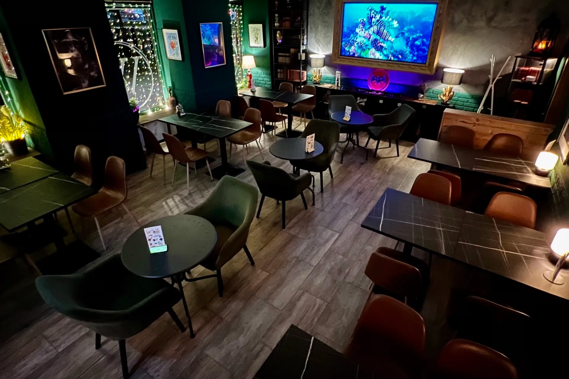 Ein gemütliches, modernes Café mit dunklen Tischen und Stühlen, Grün- und Brauntönen, Lichterketten, Wandkunst und einem großen Fernseher, der eine Unterwasserszene zeigt. Der Raum ist sanft beleuchtet und menschenleer.
