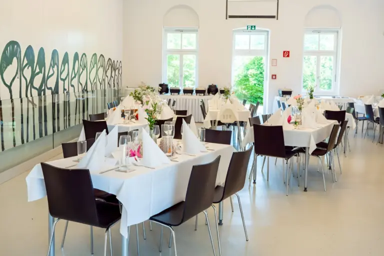 Ein heller Bankettsaal mit weißen Wänden und großen Fenstern, der für eine Veranstaltung eingerichtet ist. Die Tische sind mit weißen Tüchern, gefalteten Servietten, Gläsern und Blumensträußen eingedeckt. Dunkle Stühle umgeben jeden Tisch.