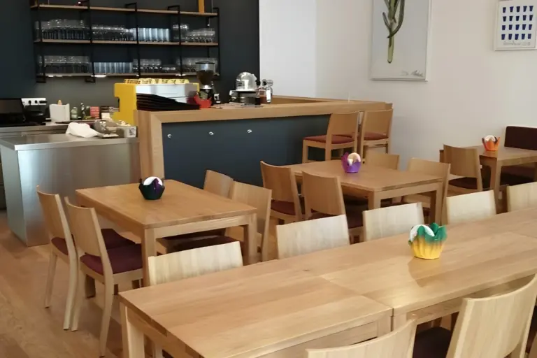 Ein kleines, modernes Café mit mehreren leeren Holztischen und -stühlen, bunten Blumentöpfen in der Mitte, einem Tresen mit Regalen mit Gläsern und Tellern und minimalem Dekor an den Wänden.