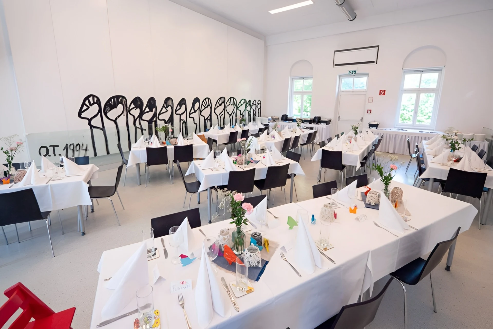 Ein hell erleuchteter Speisesaal mit weißen Wänden und großen Fenstern, der für eine Veranstaltung eingerichtet ist. Lange Tische sind mit weißen Tischtüchern, Servietten und Blumenschmuck bedeckt. Schwarze Stühle und Wandkunst sind zu sehen.