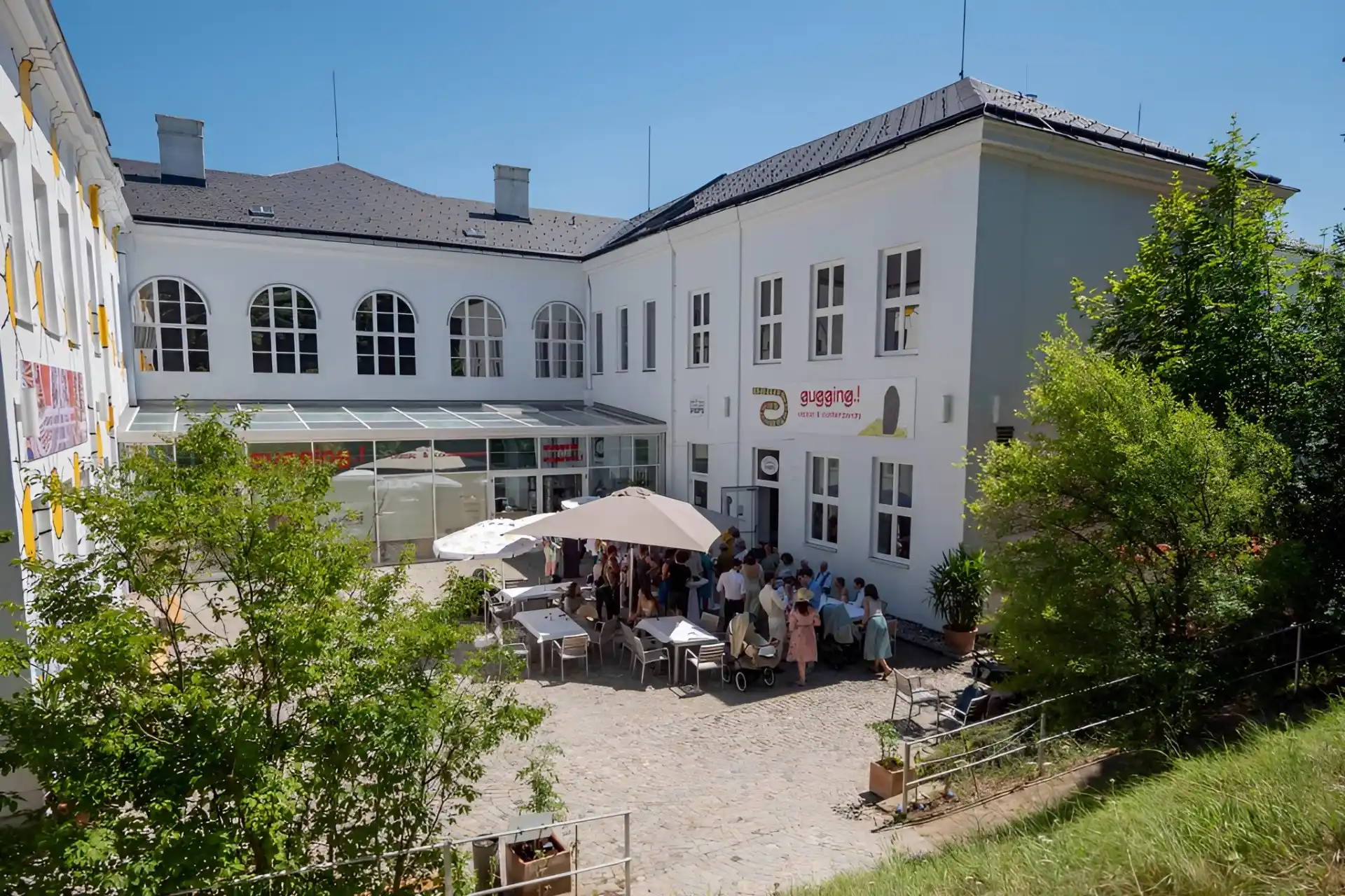 Eine Gruppe von Menschen versammelt sich an einem sonnigen Tag unter Sonnenschirmen im Innenhof eines weißen Gebäudes mit großen Fenstern. Einige Grünflächen und Bäume umgeben den Innenhof.