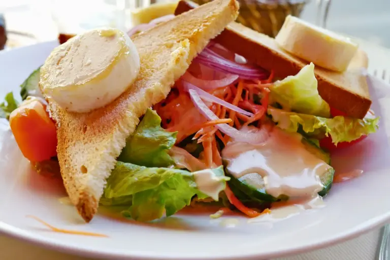 Ein Salatteller mit Salat, geschnittenen roten Zwiebeln, geraspelten Karotten, Kirschtomaten, cremigem Dressing und zwei getoasteten Brotscheiben, belegt mit runden Weichkäsescheiben.