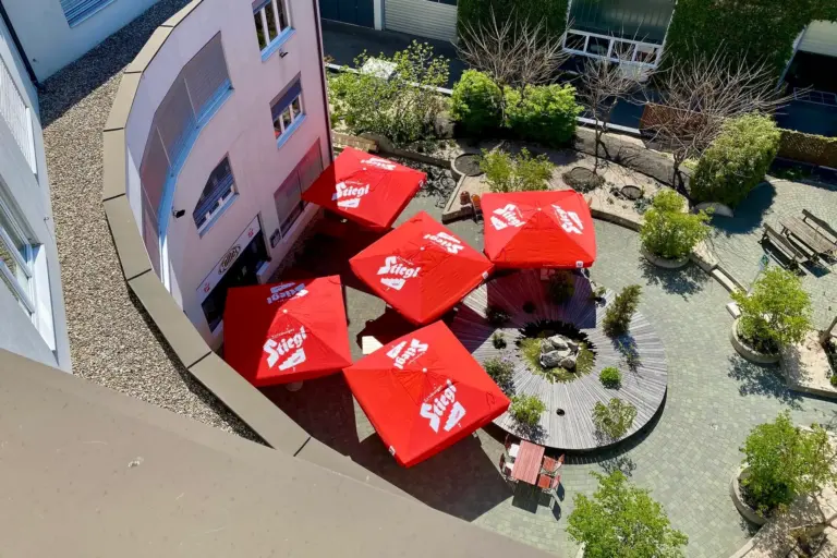 Luftaufnahme eines Innenhofs mit kreisförmigem Holzsitz und fünf roten Sonnenschirmen mit der Aufschrift Stiegl, umgeben von Grünpflanzen und Bäumen, neben einem geschwungenen Gebäude.