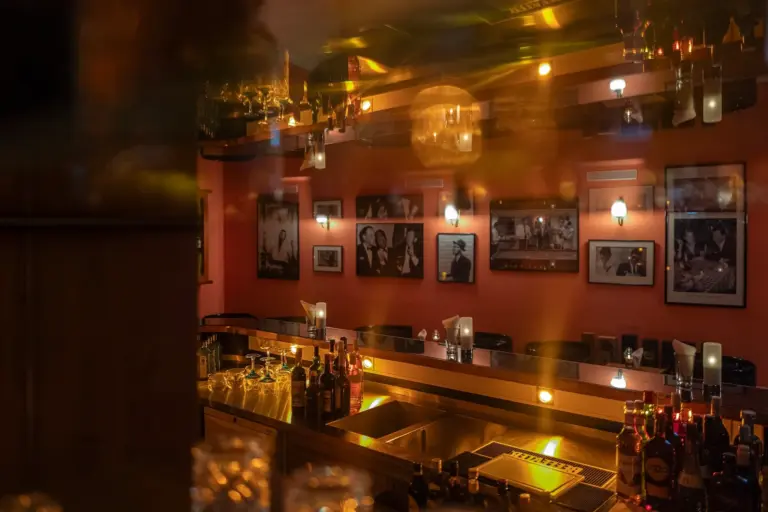 Eine schummrig beleuchtete Bar mit Flaschen auf der Theke, Barhockern, gerahmten Schwarz-Weiß-Fotos an roten Wänden und warm leuchtenden Lichtern, die sich auf den Glasflächen spiegeln.
